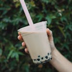Close-up of a hand holding iced milk tea with boba pearls and straw against a leafy background.