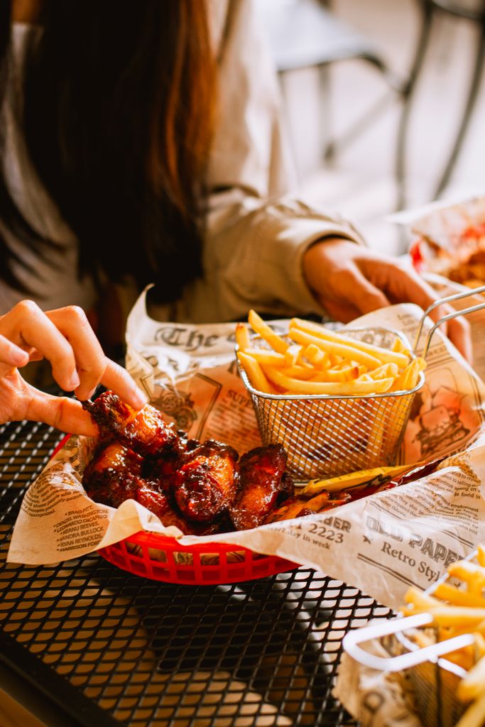 A close-up of hands enjoying a basket of BBQ chicken wings and crispy fries at a restaurant.